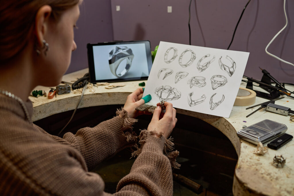 Person is working on jewelry designs using digital tablet and paper sketches at workshop table filled with tools and materials. Detailed sketches of rings are displayed